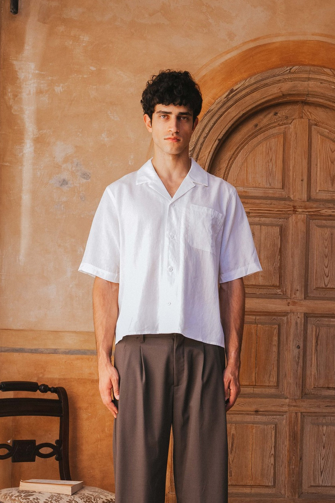 Man wearing a white shirt and brown pants standing in front of a wooden door.