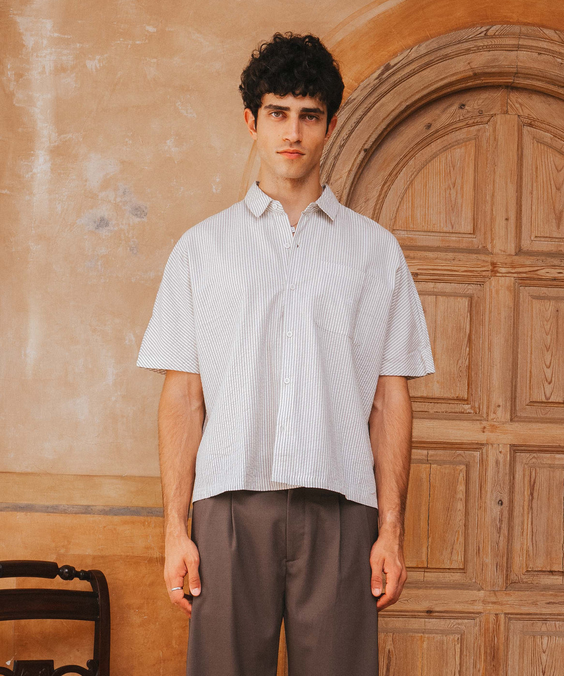 Man wearing a light-colored shirt and brown pants standing in front of a wooden door.