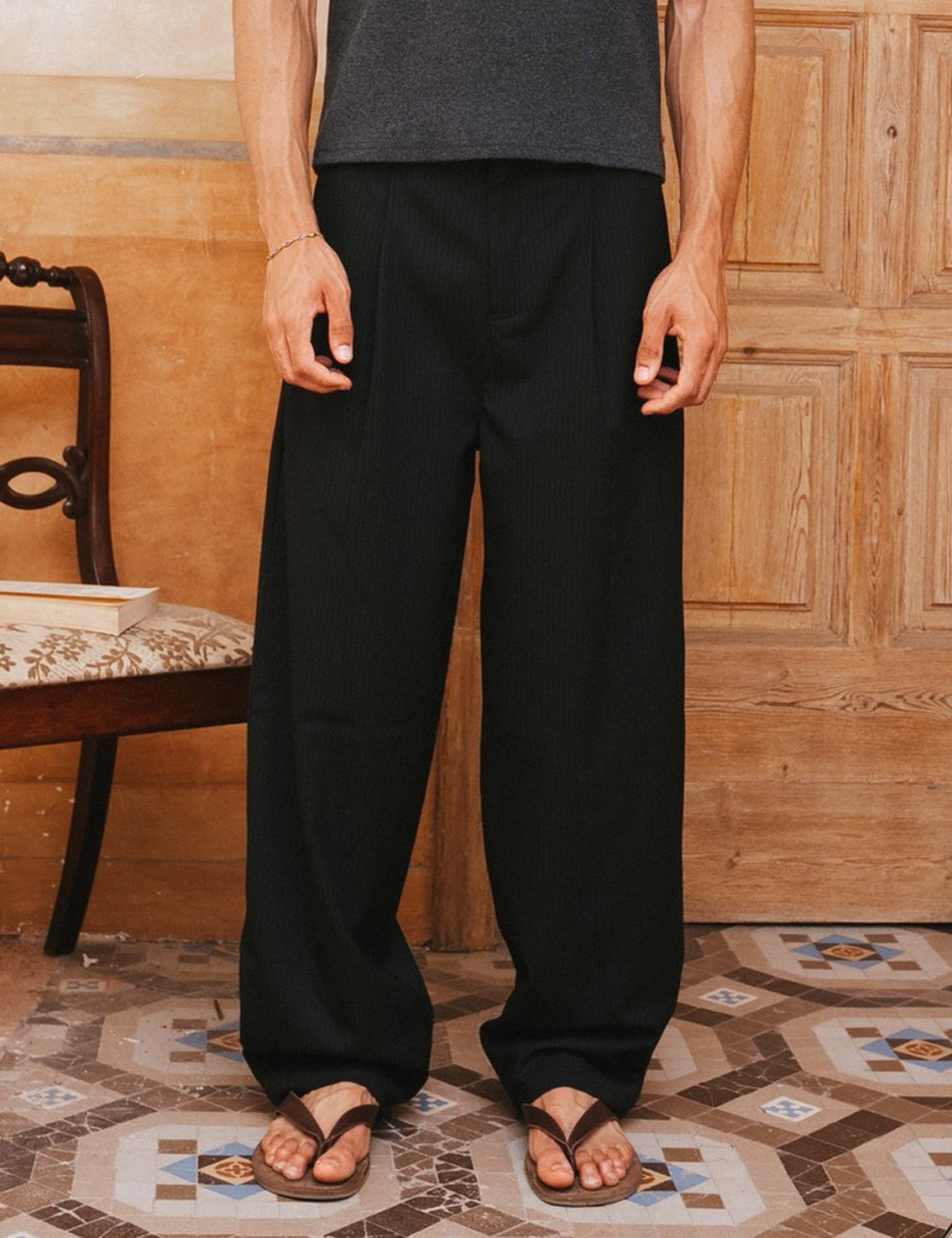 Person wearing black pants and a gray tank top standing in a room with wooden paneling and patterned floor.